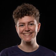 A headshot of a person with short curly hair, smiling at the camera, on a black background.