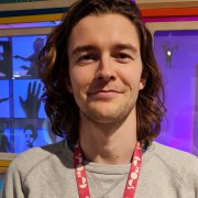 White male with long curly brown hair wearing a grey jumper. Background is a colourful display cabinet 